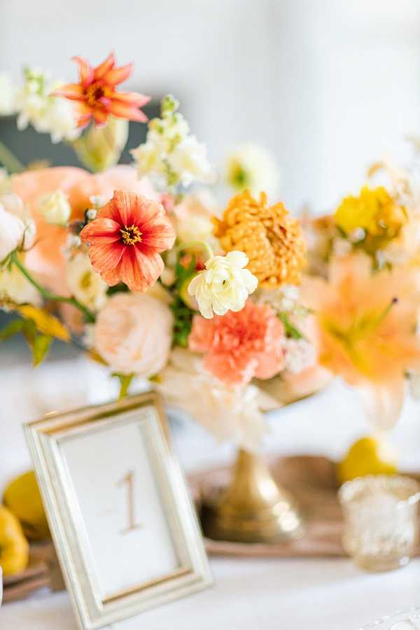Close-up detail shot of a wedding reception table centerpiece arrangement featuring coral zinnias, peach garden roses, cream ranunculus, mustard yellow marigolds, coral carnations, and cream stock flowers in gold compote vases on a gold tray. A gold-framed table number card displaying the number '1' is positioned in the foreground, and whole lemons are visible as decorative accents on the tray. The color palette is warm and citrus-toned with peach, coral, mustard yellow, and cream, styled in a garden-fresh, slightly bohemian aesthetic. The white table linen is visible beneath, and the background is softly blurred, keeping focus on the floral arrangement.