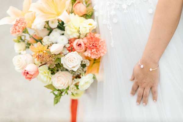 Close-up detail shot of a bride holding a bouquet, with her hand and partial torso visible. The bouquet features a mix of peach roses, coral carnations, blush garden roses, ivory ranunculus, orange lilies, small white filler flowers, and green foliage, tied with a coral ribbon. The bride wears a white dress with pearl or bead embellishment detail visible at the neckline and a delicate thin bangle bracelet with a small pearl or stone accent. The background is softly blurred, keeping focus on the flowers and the bride's hand.