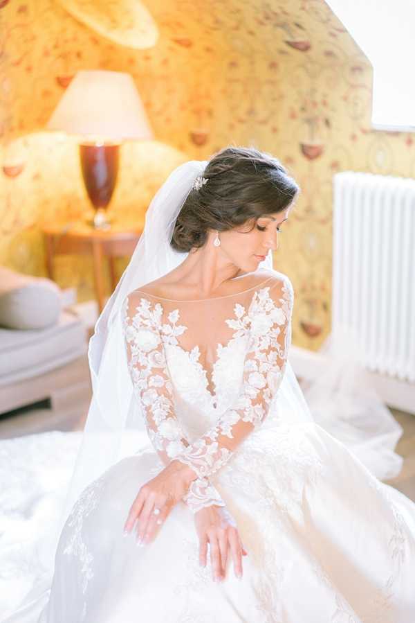 A bridal portrait taken indoors, likely in a getting-ready room of a chateau or manor house, identifiable by the yellow floral-patterned wallpaper and warm lamp light in the background. The bride is seated and looking downward, wearing a white ball gown with a sheer illusion neckline and long lace sleeves featuring a floral appliqué pattern across the bodice and arms. Her dark hair is styled in an updo with a small crystal hair accessory, and she wears a long cathedral-length veil and drop earrings. The composition is a medium portrait shot with soft, warm natural and artificial light creating a bright, airy feel.