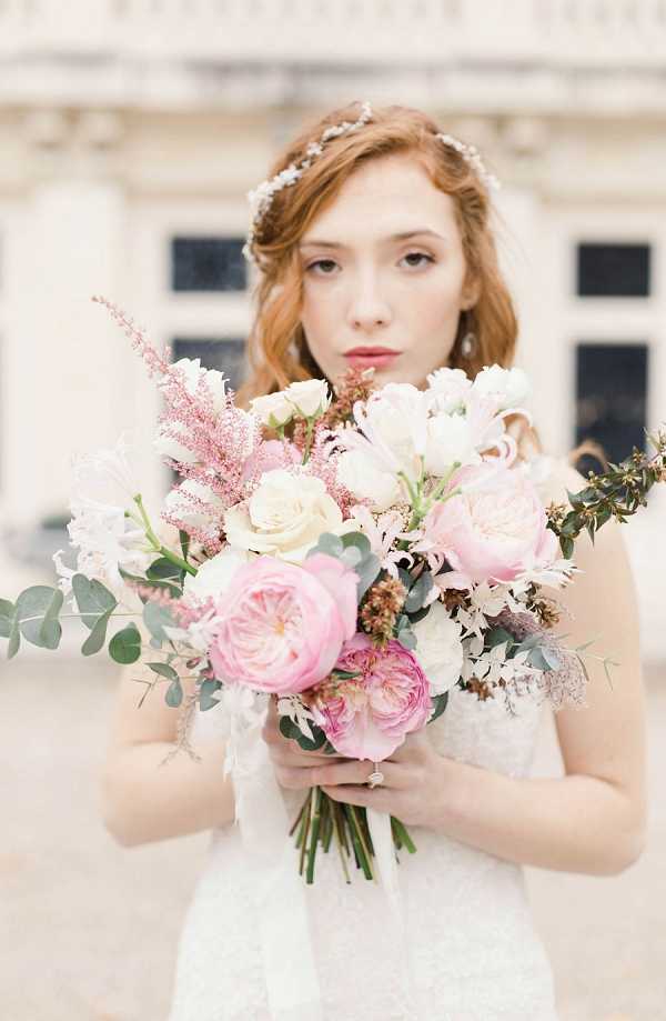 A close-up bridal portrait of a red-haired bride holding a large, loosely arranged bouquet featuring blush and hot pink garden roses, cream roses, white tulips, pink astilbe, white clematis, and eucalyptus foliage. The bride wears a white lace dress and a delicate crystal or pearl floral hair crown, with her hair styled in soft waves. The image is shot outdoors in front of what appears to be a classical stone building with large windows, slightly blurred in the background. The overall styling aesthetic is romantic and soft, with a blush and cream color palette.