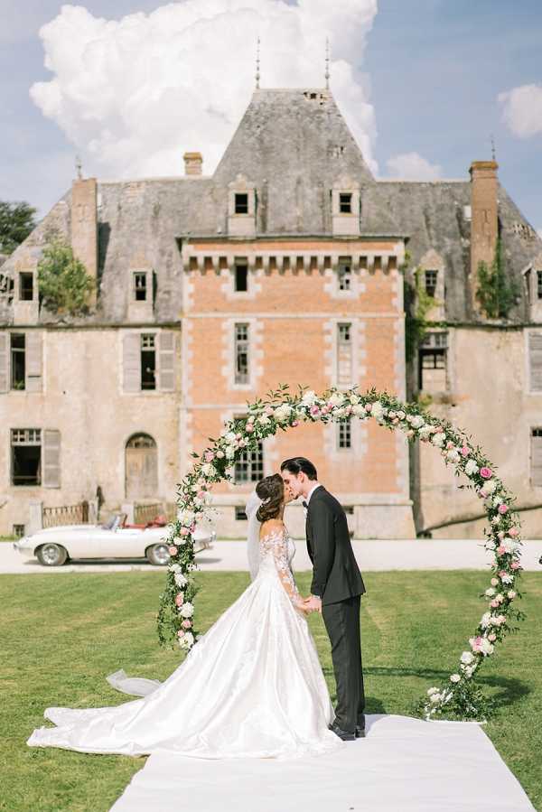 A bride and groom share a kiss under a circular floral arch during an outdoor ceremony on the grounds of a French chateau. The arch is densely decorated with blush pink and white blooms — likely garden roses and peonies — interspersed with lush greenery. The bride wears a long-sleeved, lace-bodice ball gown in ivory with a full satin skirt and an extended train, while the groom is dressed in a dark charcoal suit with a white dress shirt and bow tie. A white aisle runner leads up to the arch, and a vintage white convertible sports car is parked to the left in front of the chateau's pink brick and stone facade. The composition is a wide portrait shot with the multi-story chateau centered prominently in the background. Potential venue feature image.
