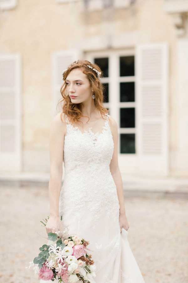 A bridal portrait taken outdoors in front of a French chateau facade with pale stone walls and white-shuttered windows. The bride stands alone, gazing to one side, wearing a fitted lace wedding gown with an illusion neckline and thin straps. Her auburn hair falls in loose waves and is accessorized with a delicate crystal or pearl hair vine. She holds a loosely arranged bouquet featuring blush and hot pink peonies, white ranunculus, dusty pink blooms, and eucalyptus foliage. The styling has a romantic, garden-inspired feel. Medium portrait crop, shot with a shallow depth of field that softly blurs the chateau background.