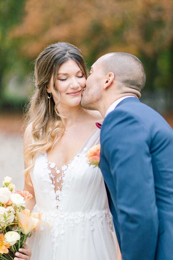 A couple portrait taken outdoors, with the groom kissing the bride on the cheek while she smiles with her eyes closed. The bride wears a white deep V-neck gown with floral lace appliqué bodice and a flowy tulle skirt, paired with gold hoop earrings and loose wavy hair. She holds a bouquet of peach tulips, ivory ranunculus, and coral blooms. The groom wears a navy blue suit with a burgundy bow tie and a peach floral boutonnière. The background is softly blurred with warm amber and gold foliage, suggesting an autumn setting. The shot is a close-up portrait with a bright, airy look.