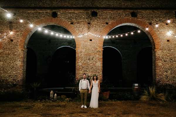 A couple poses together outdoors at night in front of a large stone and brick building featuring three prominent arched openings with dark interiors. The bride wears a flowing white gown and a floral crown, while the groom is dressed in khaki trousers, suspenders, a bow tie, and white sneakers. String lights are draped across the arches, providing warm ambient lighting against the dark sky. A wooden wine barrel is visible to the right, and a lantern sits on the ground to the left, suggesting a rustic or boho styling theme; this is a wide couple portrait shot taken at night.