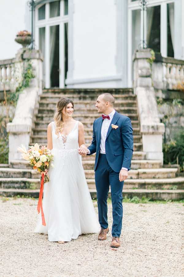 A couple portrait taken outdoors on a gravel path in front of a chateau with ornate stone balustrades and a grand staircase. The bride wears a white sleeveless gown with a deep V-neckline and lace bodice detail, holding a loose bouquet of autumn-toned flowers including orange and yellow blooms with pampas grass accents and a coral ribbon tie. The groom wears a fitted navy blue suit with a burgundy bow tie, white dress shirt, and a yellow-orange boutonniere. The couple holds hands and looks at each other, smiling. The overall styling reflects a warm autumnal palette with a classic-meets-modern aesthetic. Medium portrait shot with soft natural light.