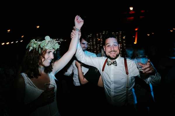 The couple is dancing energetically on the dance floor during the evening reception, with several guests visible in the background. The bride wears a white lace dress and a greenery crown made of eucalyptus-style leaves in her hair, while the groom is dressed in a white shirt with brown leather suspenders and a patterned bow tie. Both are holding drinks and have their joined hands raised in the air, laughing. The setting is an indoor venue lit with warm amber uplighting and string lights in the background, creating a dark, festive atmosphere consistent with a boho rustic styling theme. The shot is a candid mid-range portrait capturing the lively mood of the reception.
