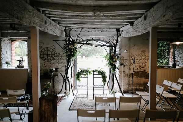 A wide shot of an indoor ceremony setup inside a rustic, industrial-style space with whitewashed exposed brick walls, weathered wooden ceiling beams, and a concrete floor. The ceremony arch is constructed from dark twisted branches, decorated with lush greenery and small clusters of cream and blush flowers, and is positioned in an open doorway that frames an outdoor garden beyond. White and natural wood folding chairs are arranged in rows on either side of the aisle, with a patterned vintage-style rug placed at the altar area. The decor style is rustic-organic, mixing raw architectural elements with botanical details, and no guests or couple are present in the image. Potential venue feature image.