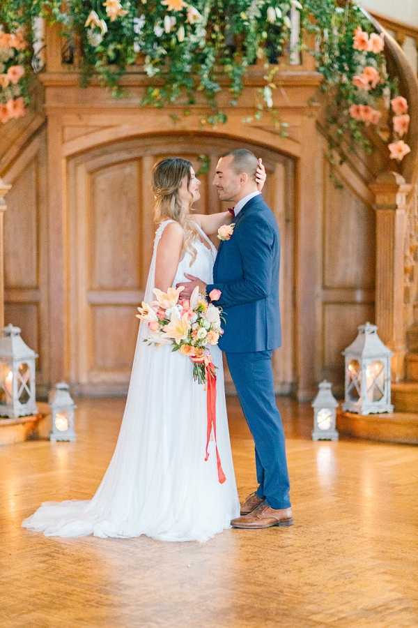 A bride and groom share an intimate moment on a polished wooden dance floor inside a venue with carved wooden paneling and arched doorways. The bride wears a white sleeveless A-line gown with a V-neckline and flowing skirt, holding a bouquet of coral, orange, and peach blooms including lilies and roses tied with a red ribbon streamer. The groom wears a navy blue suit with brown leather oxford shoes and a coral boutonniere. Overhead, a lush hanging floral installation features trailing greenery and peach-coral flowers, and white lanterns with candles are arranged along the base of the wooden paneling on both sides. The portrait is a mid-length couple shot with soft, bright natural-style lighting and a warm, romantic color palette centered on coral and peach tones.
