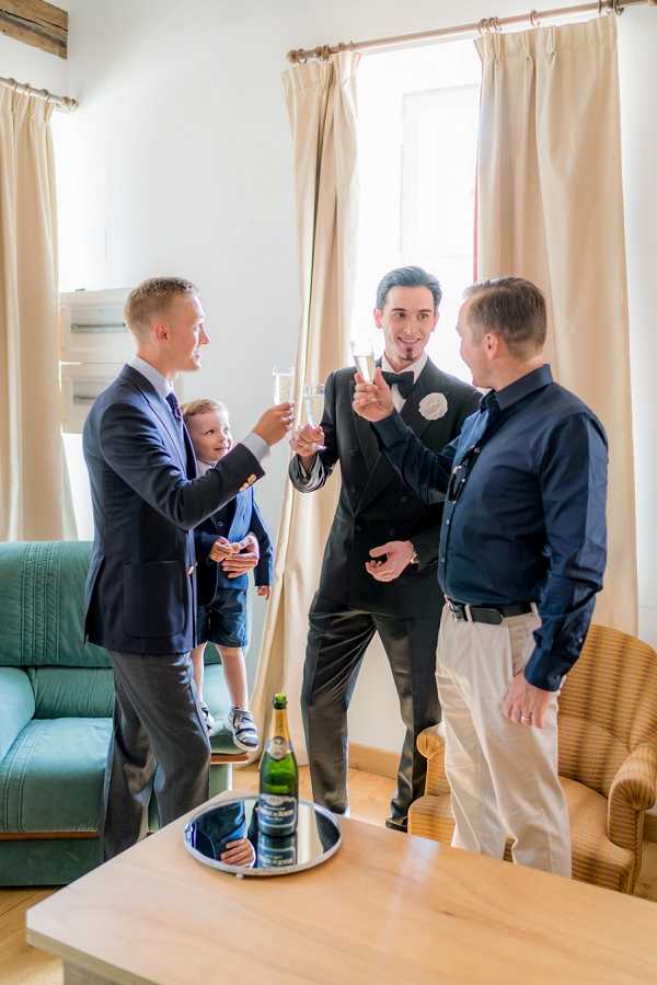 A getting-ready moment captured indoors in a simply furnished room with cream curtains, a teal sofa, and a wooden coffee table. Three men and a young boy are sharing a champagne toast — the groom is dressed in a black tuxedo with a black bow tie and a white boutonniere, while a groomsman wears a navy suit, and an older man in a navy shirt and khaki trousers joins in. A young boy in a navy outfit stands beside the groomsman. A bottle of champagne sits on the floor near a silver tray on the coffee table that holds folded navy accessories. The shot is a candid medium-wide portrait taken from a slightly low angle.