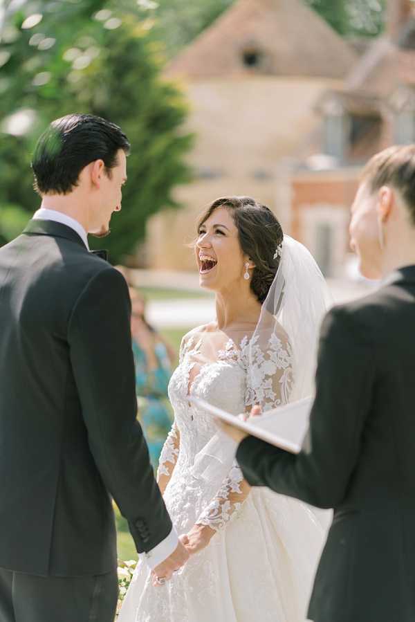 An outdoor wedding ceremony capturing a candid moment of the bride laughing openly while holding hands with the groom, with an officiant visible in the foreground right holding a booklet. The bride wears a long-sleeve ivory lace gown with floral appliqué detailing and a cathedral-length veil, accessorized with drop earrings and a hair piece; the groom is dressed in a black tuxedo with a bow tie. The ceremony takes place in a sunny garden setting with a stone and brick chateau building visible in the soft-focus background, along with a small number of seated guests. The image is a medium portrait shot with a shallow depth of field, emphasizing the bride's joyful expression.
