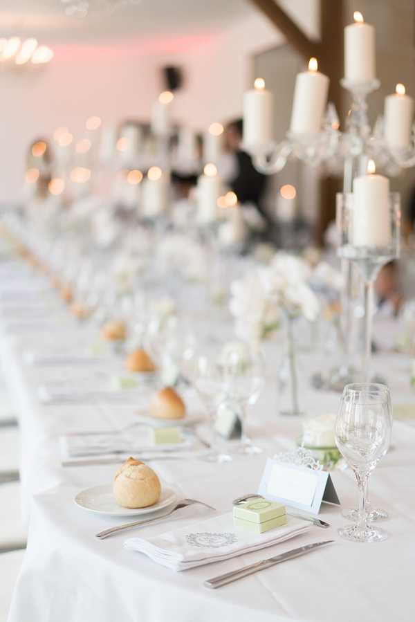 A detail shot of a long reception banquet table set for a wedding dinner in an indoor venue with exposed wooden beams and soft pink ambient lighting. The table is dressed in a white linen cloth with place settings featuring silver cutlery, crystal wine glasses, white monogrammed napkins, small green favour boxes, and white place cards. Individual bread rolls sit on side plates at each setting. Tall crystal candelabras holding white pillar candles run along the centre of the table, alternating with low arrangements of white flowers, likely roses and peonies. The table stretches into the background where guests can be seen seated, creating a strong sense of depth. The overall decor palette is all-white and crystal with classic, formal styling.