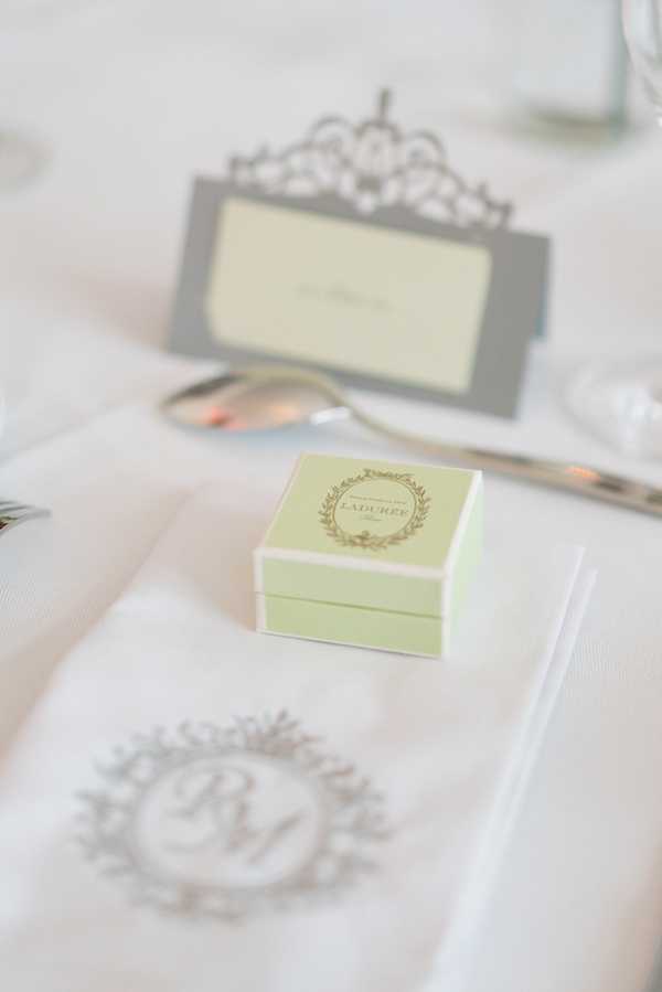 A close-up detail shot of a wedding reception place setting featuring a pale mint green Ladurée favor box centered on a white linen napkin embroidered with a monogrammed crest in silver-grey thread. Silver cutlery is visible alongside the napkin, and an ornate silver filigree place card holder with a cream card sits slightly out of focus in the background. The overall decor palette is white, silver-grey, and mint green, suggesting a classic French-inspired styling theme.