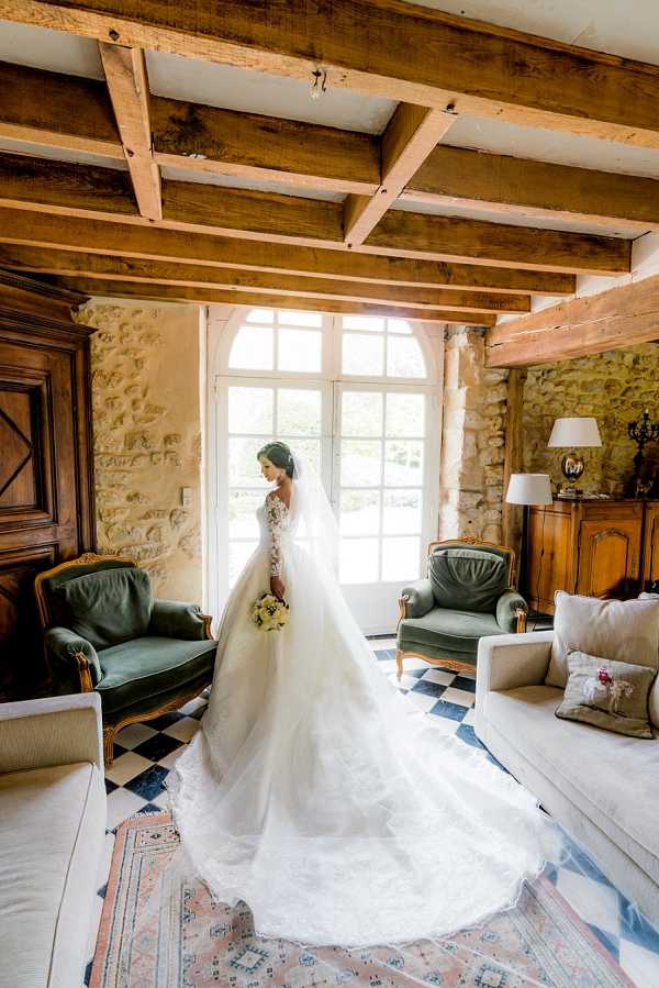 A bridal portrait taken indoors in what appears to be a chateau salon or sitting room, with the bride standing in front of a large arched French window that backlit the scene with natural light. The bride wears a full-volume ball gown with long lace sleeves and an exceptionally long cathedral train spread across the black-and-white checkered tile floor, and she holds a small bouquet of cream and pale yellow flowers. The room features exposed wooden ceiling beams, stone walls, green velvet Louis XV-style armchairs with gilt frames, cream upholstered sofas, a vintage Persian-style area rug in muted pink and blue tones, and a wooden antique sideboard with a table lamp to the right. The composition is a full-length portrait shot from a slight distance, capturing the full spread of the train, with the bride facing away toward the window in a three-quarter profile pose. Potential venue feature image.