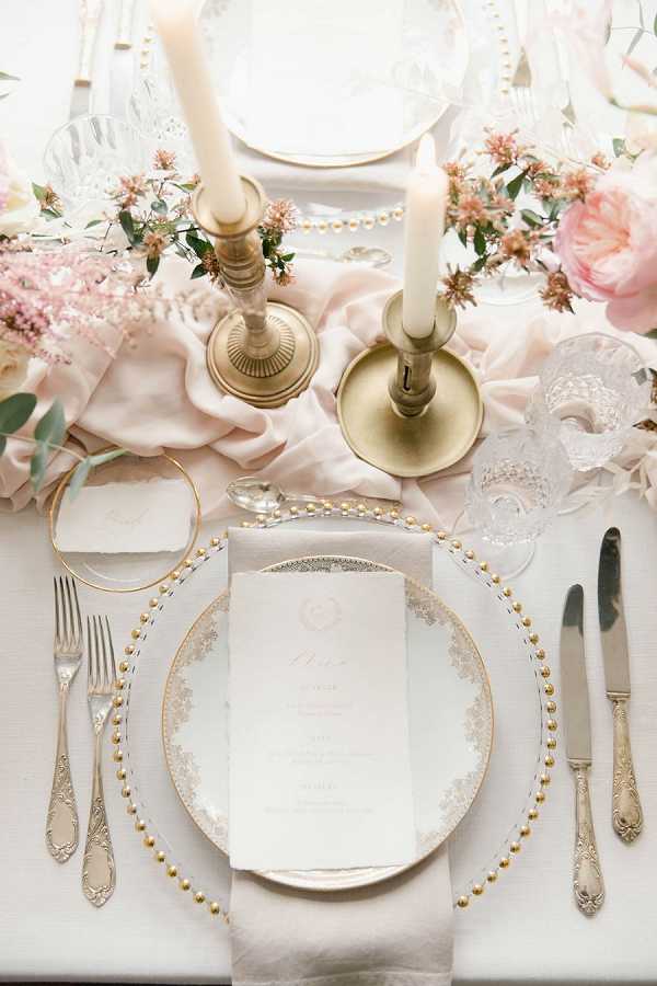 Close-up detail shot of a wedding reception table setting styled in a classic French romantic palette of blush pink and gold. The place setting features a gold beaded charger plate topped with a white lace-patterned china plate and a handwritten menu card with a small floral motif; antique-style silver cutlery is arranged on either side. Two brass candlestick holders with ivory taper candles anchor the center, alongside a blush pink silk table runner draped loosely across a white linen tablecloth. Floral arrangements of blush pink peonies, dusty pink astrantia, and small dried pink blooms with eucalyptus foliage are visible at the edges of the frame, and cut crystal glassware and a gold ring place card holder complete the setting.