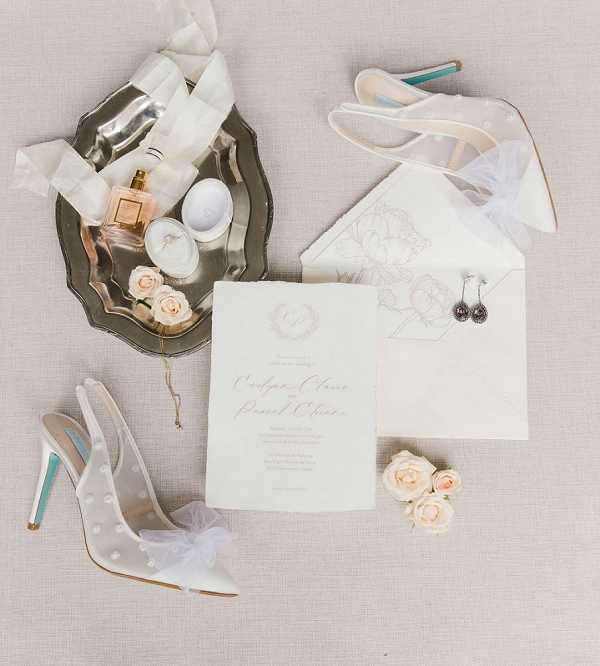 A flat lay composition of bridal details arranged on a neutral linen-toned surface. The items include a white wedding invitation suite with a monogram crest and an envelope lined with a delicate floral illustration, a silver tray holding a gold perfume bottle, a small white ring box, silk ribbon, and several blush pink roses, a pair of white pointed-toe heels with pearl embellishments and teal-colored stiletto heels, a second pair of white strappy heels partially visible at the top, small drop earrings, and loose blush pink roses scattered around the arrangement. The overall color palette is white, ivory, blush, and gold with teal accents on the shoes.