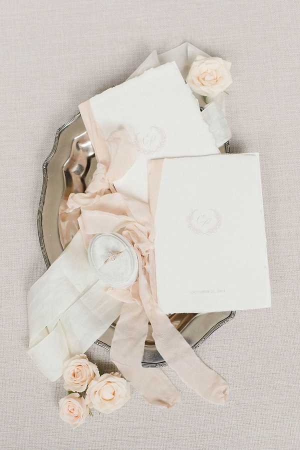 A flat lay detail shot of a wedding stationery and accessories styled arrangement on a linen-toned background. Two ivory deckle-edged invitation suite booklets with an embossed monogram crest are displayed on an ornate silver tray, tied with blush silk ribbon. A small round velvet ring box in pale blue-grey holds a diamond engagement ring, and several blush spray roses are scattered around the tray as styling accents. The overall palette is ivory, blush, and silver with a classic, refined paper goods aesthetic.