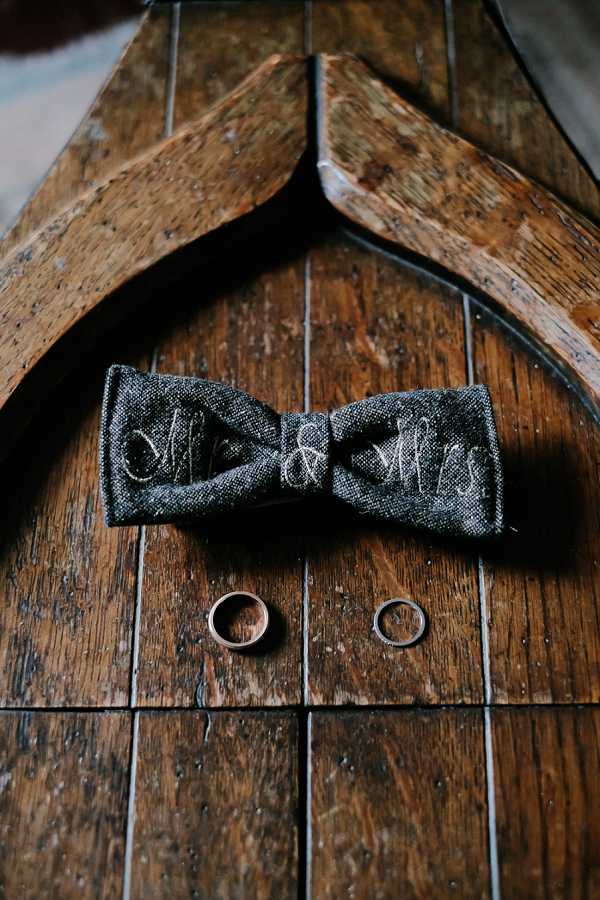 A close-up detail flat lay shot of wedding accessories arranged on a worn wooden surface. A charcoal grey textured bow tie embroidered with 'Mr & Mrs' in silver script is positioned beneath a rustic wooden hanger, with two wedding bands placed below it — one wider gold-toned band and one slimmer, more delicate band. The styling is rustic with a dark, moody tone.