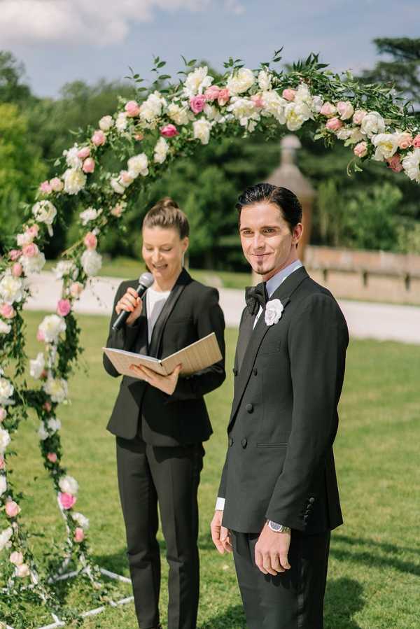 An outdoor wedding ceremony in a garden setting, showing the groom standing at the altar and looking toward the camera while a female officiant reads from a ceremony booklet into a microphone behind him. The groom wears a black double-breasted suit with a black bow tie, a light blue dress shirt, and a white floral boutonniere. The officiant is dressed in a black pantsuit. They stand beneath a circular floral arch densely covered with ivory peonies, blush pink roses, and lush greenery. The composition is a mid-shot portrait capturing both figures with the arch framing the scene.
