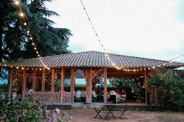 Wide shot of an outdoor wedding reception at dusk, taking place inside an open-sided rustic wooden pavilion with a terracotta tile roof. Strings of warm Edison bulb fairy lights are draped across the structure and extend outward from the roofline, creating a warm glow against the darkening sky. Guests, approximately 15–20 visible, are seated at tables inside the pavilion, dressed in formal attire including white shirts and dark clothing. Small bistro tables and folding chairs are also placed outside the pavilion in the foreground. The overall styling is rustic with natural wood construction and warm string lighting. Potential venue feature image.