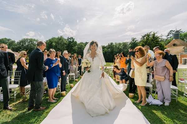 The bride walks down the aisle during an outdoor ceremony, flanked by approximately 30-40 seated and standing guests on either side. She wears a long-sleeved ivory ball gown with a full skirt and a cathedral-length veil, carrying a compact bouquet of white and cream flowers. The ceremony is set on a manicured lawn with white Chiavari chairs arranged in rows, and a white aisle runner leads toward the altar. A brick outbuilding is visible in the background, suggesting a chateau or estate venue. The composition is a wide mid-shot taken from ground level looking toward the bride, capturing the full scene in bright midday sunlight.
