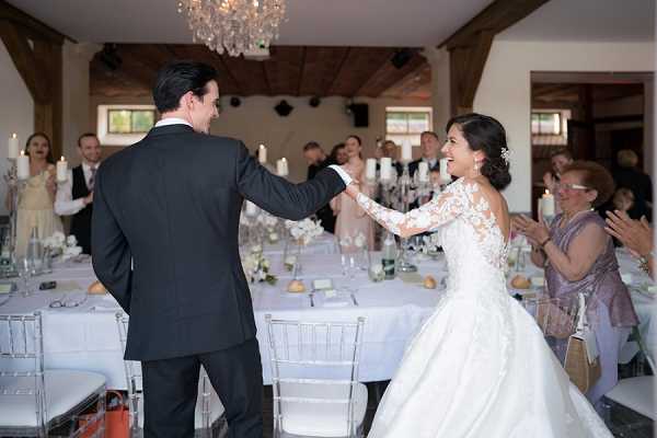 The bride and groom are making their entrance into the wedding reception hall, holding hands and facing each other as guests applaud around them. The indoor venue features exposed wooden beams, a crystal chandelier overhead, and tall white pillar candles as centerpieces on tables dressed with white linens and silver chiavari chairs. The bride wears a long-sleeve lace ballgown with an illusion bodice and has her hair pinned up with a floral accessory, while the groom wears a dark charcoal suit. Approximately 15-20 guests are visible in the background, dressed in formal attire. The image is a wide shot capturing the full scene from behind the groom's left shoulder.