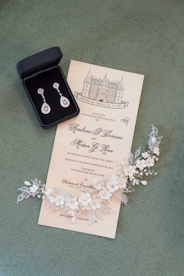 A flat lay detail shot of wedding stationery and accessories arranged on a green textured surface. The centerpiece is a cream-colored wedding invitation for Roselina S. Doneva and Morten G. Lowe, featuring a hand-drawn illustration of Château de Courselles Le Roy at the top and script typography throughout, noting the ceremony date of Tuesday 04-06-2019 at 16:00. A black jewelry box containing a pair of crystal drop earrings with teardrop-shaped cubic zirconia settings sits in the upper left corner, and a delicate ivory floral hair comb with small white blossoms and silver beaded accents is partially overlapping the lower right of the invitation.