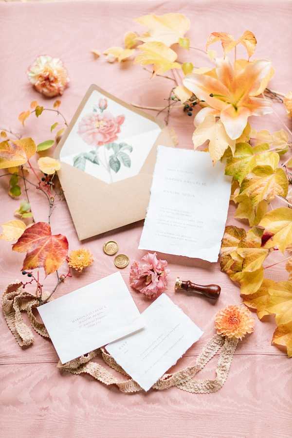 A flat lay detail shot of a wedding stationery suite arranged on a dusty pink linen surface. The suite includes a main invitation card and RSVP card with deckled edges in white with serif typography, a kraft paper envelope lined with a botanical blush pink rose illustration, two gold wax seals, and a wooden wax stamp. The arrangement is styled with scattered autumnal foliage in amber, yellow, and rust tones, small pink carnations, a peach dahlia, and a length of cream lace ribbon. The overall styling palette is blush, warm gold, and terracotta, suggesting a romantic autumn wedding aesthetic.