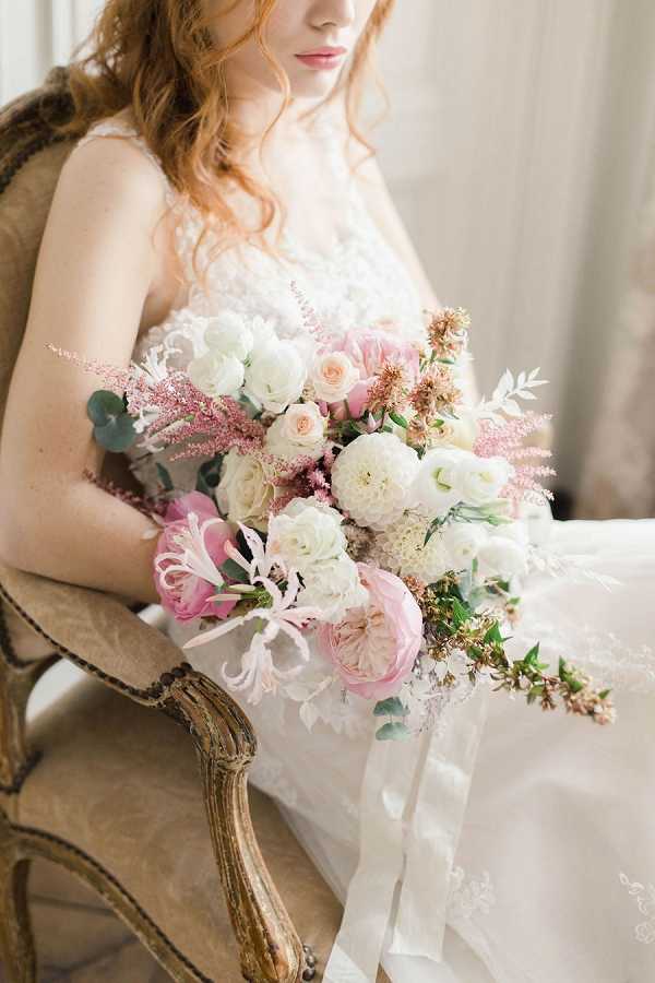 A bridal portrait shot indoors, with a bride seated in an antique gilded French-style armchair holding a large, loosely arranged bouquet. The bride wears a white lace wedding dress and has loose auburn waves; her face is partially cropped out of the frame. The bouquet features blush and deep pink garden roses, white ranunculus, white dahlias, pink astilbe, dusty rose dried florals, pale pink honeysuckle-style blooms, and eucalyptus foliage, tied with long trailing ivory satin ribbons. The overall styling is romantic and soft, with a pale neutral interior backdrop and a classic French aesthetic. This is a close-to-medium portrait composition focused on the bouquet and dress detail.