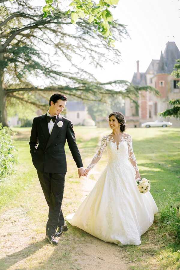 A couple portrait taken outdoors on the grounds of a French chateau, with the historic brick and stone manor house visible in the soft-focus background. The bride wears an ivory ball gown with sheer lace long sleeves and floral appliqué detailing, carrying a small bouquet of white blooms, while the groom wears a black double-breasted tuxedo with a black bow tie and a white floral boutonnière. The two are walking hand-in-hand along a gravel path, both smiling and looking at each other. The shot is a medium full-length portrait taken in bright natural daylight with a classic, formal styling aesthetic.