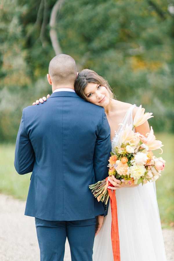 A couple portrait taken outdoors on a gravel path with a softly blurred green tree backdrop. The bride leans her head against the groom's shoulder and looks back toward the camera, wearing a white V-neck gown with delicate embellishment at the neckline. The groom faces away from the camera, dressed in a navy blue suit. The bride holds a large, loosely arranged bouquet featuring peach lilies, orange dahlias, ivory blooms, yellow flowers, and foliage, tied with a trailing burnt orange ribbon. The composition is a mid-length portrait shot from behind the couple, with the bride's face as the focal point.