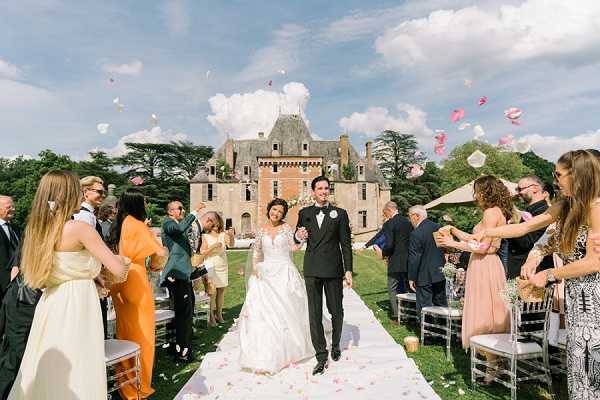 The bride and groom walk back up the aisle together following their outdoor wedding ceremony, with approximately 30 guests lining either side tossing pink and white flower petals into the air. The bride wears a long-sleeved lace ball gown in ivory, and the groom is dressed in a black tuxedo with a white boutonniere. The ceremony took place on a white aisle runner lined with ghost chairs on a lawn directly in front of a large French chateau with a distinctive slate-roofed tower and red-brick facade. Guests are dressed in a variety of colors including blush, orange, and yellow. The shot is a wide, eye-level portrait framing the couple centered between the two rows of guests with the chateau prominently visible in the background. Potential venue feature image.