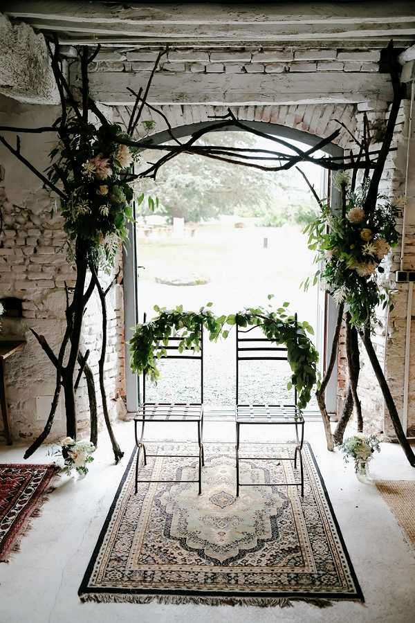 A ceremony altar setup inside a rustic venue with exposed whitewashed brick walls, featuring two black wrought-iron chairs placed on a vintage Persian-style rug in cream and navy tones. The arch is constructed from large, dark bare branches arranged dramatically around an open doorway, adorned with clusters of greenery and cream and blush flowers — including what appear to be dahlias, chrysanthemums, and foliage — at the base of each side. The two chairs are draped with lush garlands of mixed green foliage. Small floral arrangements in white and cream are placed at floor level near the base of the branch installation, and a second red-toned rug is partially visible to the left. The overall styling is organic and boho with a naturalistic, woodland-inspired aesthetic. Wide shot, no people present. Potential venue feature image.