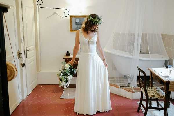 A bride is photographed getting ready in what appears to be a room inside a French chateau or country house, looking down at her bouquet in a candid moment. She wears a flowing white chiffon dress with a V-neckline and lightly structured bodice, and a full green leafy floral crown in her upswept hair. Her bouquet features white blooms, greenery, and fern fronds in a loose, organic arrangement. The room has terracotta tile floors, white walls, a freestanding white clawfoot bathtub partially visible behind a sheer white canopy, a rustic wooden side table, and a gold-framed mirror on the wall, suggesting a boho-rustic styling theme. The shot is a full-length portrait taken indoors with natural light.