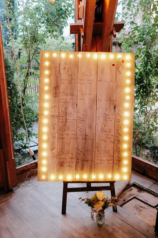 A close-up shot of a wedding seating chart displayed on a rustic wooden pallet board edged with warm-white marquee bulb lights, mounted on a wooden easel. The guest names and table numbers are handwritten directly onto the natural wood planks in what appears to be a casual, informal style. A small glass vase holding dried yellow flowers sits at the base of the easel. The setting appears to be an indoor-outdoor wooden structure with greenery and a deck visible through large windows or open walls in the background, suggesting a rustic or boho styling theme.
