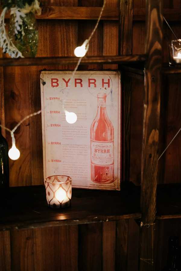 A close-up detail shot of a rustic bar or reception area decorated with a vintage Byrrh aperitif tin advertising sign in red and cream tones, propped against a dark wooden slatted wall. A faceted glass votive candle holder glows warmly in the foreground left, and a string of globe fairy lights runs across the frame adding warm ambient light. The overall styling is rustic and vintage-inspired, with the antique French liquor signage serving as a decorative prop. A dark green glass bottle is partially visible on the left edge of the frame.