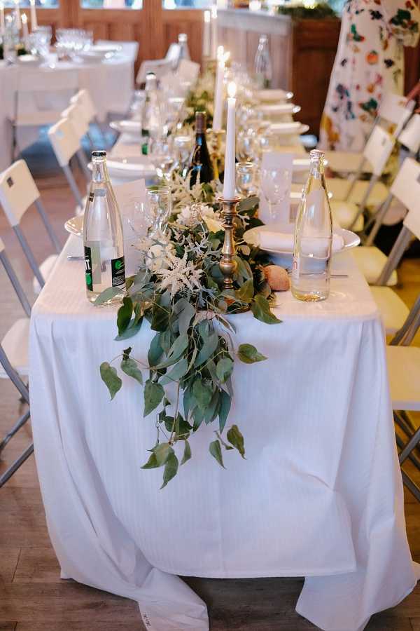 Close-up detail shot of a long reception dining table set up indoors in a wood-paneled room with warm lighting. The table is covered in a white linen tablecloth and features a lush greenery runner made of large-leafed branches and small white astilbe-style blooms cascading off the end of the table. Brass candlestick holders with tall white taper candles run along the center, accompanied by glass water bottles, wine bottles, crystal stemware, and white plates with folded napkins. White folding chairs line both sides of the table, with additional tables visible in the background. The overall decor palette is white and green with brass accents, leaning toward a natural, organic styling approach.