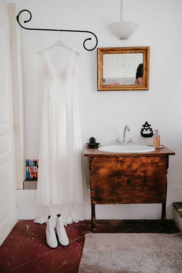 A getting-ready detail shot showing a white bridal gown hanging from a decorative black iron wall hook, with a pair of white ankle boots placed on the floor beneath it. The dress features a deep V-neckline with lace detailing at the bodice and a flowing chiffon skirt. The setting is an indoor room with a rustic-style wooden vanity unit repurposed as a bathroom sink, topped with a white basin and chrome faucet, with a gold-framed mirror mounted above it. A neutral woven rug sits on worn terracotta tile flooring, and a pendant light is visible overhead. The overall styling suggests a boho aesthetic, with the white boots as a deliberate unconventional bridal accessory choice. Wide portrait composition.