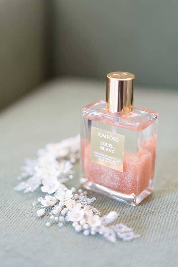 A close-up detail shot of bridal getting-ready accessories arranged on a pale sage green upholstered surface. A Tom Ford Soleil Blanc Shimmering Body Oil bottle with a rose gold cap and pink-tinted shimmer liquid is the central focus, surrounded by a delicate bridal hair accessory featuring small white fabric flowers, pearl-like beads, and silver wire detailing, consistent with a hair vine or comb style. The styling reflects a classic, polished bridal aesthetic with a rose gold and white color palette.