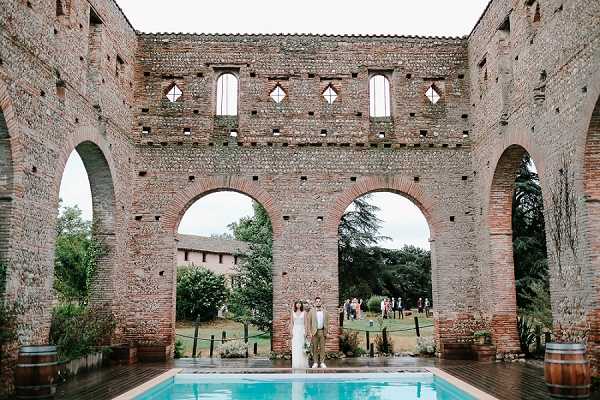 A couple stands together beneath the central arch of a large roofless brick ruin structure, positioned at the far end of a rectangular turquoise swimming pool. The bride wears a white dress and the groom is dressed in a tan or olive suit. In the background through the arches, a group of approximately 15–20 guests mingles on a lawn. Wooden wine barrels are placed on either side of the pool. The wide shot frames the couple symmetrically within the towering red-brick arcade, suggesting a rustic, historic venue setting. Potential venue feature image.