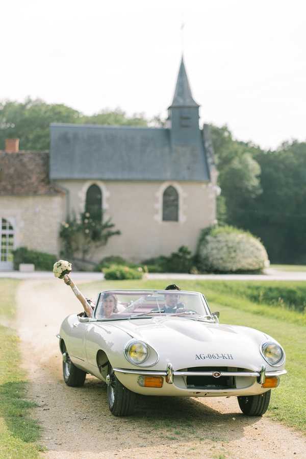 A bride and groom are driving away from a small stone chapel in a cream vintage Jaguar E-Type convertible, captured in a wide portrait-orientation shot. The bride is visible in the passenger seat with her arm raised, holding a white and green bouquet aloft in celebration, while the groom drives along a gravel driveway. The chapel in the background is a classic French country-style building with gothic arched windows, a grey slate roof, and a pointed steeple. The overall styling is classic and romantic, with the cream convertible serving as an intentional decor choice that complements the historic setting. Potential venue feature image.