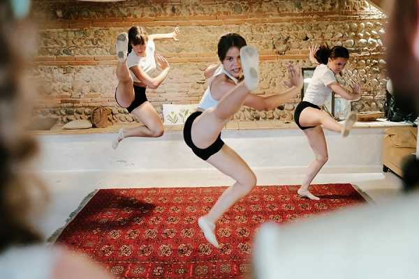 This image does not appear to be a wedding photograph. It shows three young women or teenagers performing dance or martial arts moves indoors, wearing white tops and black shorts with light-colored ballet or dance shoes. They are captured mid-movement with legs raised in a kicking or dance pose. The setting is a rustic interior space with exposed stone walls and a red patterned rug on the floor. The shot is a wide action photograph taken at a low angle, with slight foreground blur from an object partially obscuring the lens.