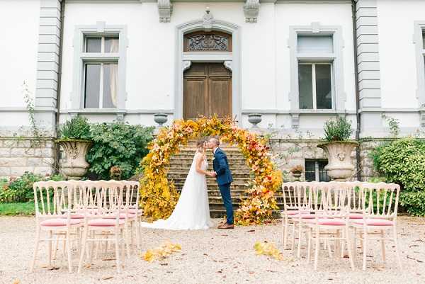 An outdoor civil or symbolic ceremony is taking place in front of a white French manor house, where the bride and groom stand beneath a large circular floral arch positioned at the base of stone steps leading to a grand wooden door. The arch is densely covered in autumn foliage and blooms in shades of deep yellow, burnt orange, rust, and burgundy. The bride wears a white tulle ballgown and the groom wears a navy blue suit; they are facing each other and appear to be exchanging vows or sharing a kiss. Rows of white Chiavari chairs with blush pink cushions are arranged on either side of a gravel aisle, with no guests visible, suggesting this may be a styled shoot or the image was taken before or after the ceremony. The overall styling is an autumnal palette with a classic French estate backdrop. Wide portrait-style shot capturing both the couple and the full venue facade. Potential venue feature image.