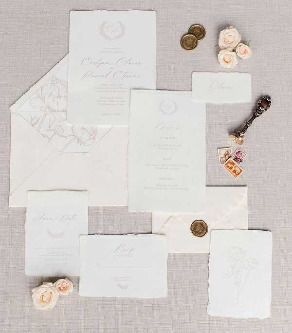 A flat lay of a complete wedding stationery suite arranged on a light grey linen background. The suite includes a main invitation with calligraphy script, an envelope with a delicate floral line-art liner, a menu card, a save the date, an RSVP card, a place card, and a loose illustrated card featuring botanical line art. Styling accents include small blush spray roses, antique gold wax coins, vintage postage stamps, an ornate wax seal stamp tool, and a gold wax seal on one envelope. The paper pieces feature deckled torn edges throughout, and the print palette is soft warm grey and gold on ivory, giving the suite a classic, refined aesthetic. The composition is a styled overhead flat lay detail shot.