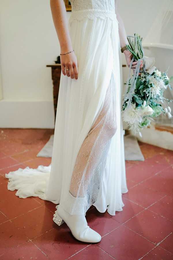 A close-up detail portrait of a bride from the waist down, standing on terracotta tile flooring inside what appears to be a French venue. She is wearing a white chiffon wedding dress with a high slit revealing a lace underskirt layer beneath, along with white block-heel ankle boots embellished with pearl details. She holds a bouquet featuring white dahlias, eucalyptus, and green foliage with a pale grey ribbon wrap. Her accessories include a delicate bracelet and a simple ring. The styling is modern with a bohemian edge, mixing bridal lace with unconventional white boots.