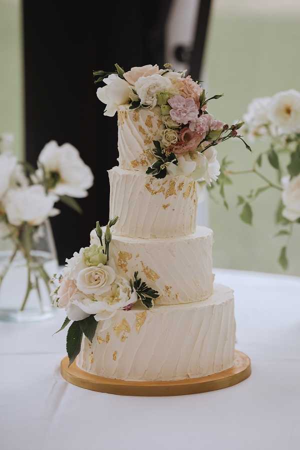 Close-up detail shot of a four-tier wedding cake displayed on a round wooden board atop a white linen-covered table. The cake is frosted in ivory buttercream with a textured vertical stripe pattern and decorated with scattered gold leaf accents across all tiers. Fresh floral clusters adorn the cake at two points — the top tier features a cascading arrangement of blush peonies, mauve roses, white lisianthus, and greenery, while the lower tiers carry a smaller cluster of cream garden roses, white lisianthus buds, and foliage. The background shows soft-focus bud vases holding white roses and greenery against a pale sage green wall, suggesting an indoor reception setting with a classic, garden-inspired decor palette.