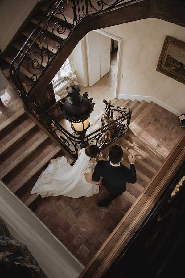 An aerial shot looking straight down at a couple standing at the base of a grand interior staircase inside what appears to be a French chateau or manor house. The bride wears a full-length ivory gown with a long trailing veil, and the groom is dressed in a dark navy or black suit; they are holding hands and facing each other. The staircase features ornate wrought-iron banisters with scrollwork details and dark wood handrails, with a large antique lantern-style pendant light hanging at the center of the stairwell. The flooring at the base is terracotta tile in a parquet-style pattern, and a gilt-framed painting is visible on the wall in the background. The composition is a dramatic overhead portrait that highlights both the couple and the architectural details of the venue interior. Potential venue feature image.