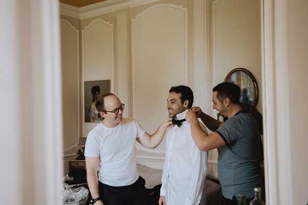A getting-ready moment showing the groom being helped with his black bow tie by two men, likely groomsmen or family members, inside a room with cream-painted wood-panelled walls and a decorative mirror in the background. The groom is wearing a white dress shirt and smiling, while one helper wears a white t-shirt and glasses and the other wears a grey t-shirt. The shot is framed through a doorway, giving a candid, voyeuristic composition, captured as a medium portrait in natural indoor light.