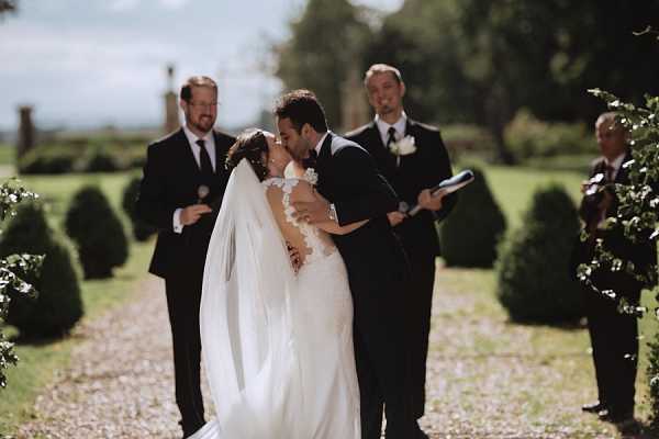 The bride and groom share their first kiss during an outdoor ceremony on a gravel path lined with trimmed topiary shrubs. The bride wears a fitted white gown with lace appliqué detailing on the bodice and a long cathedral-length veil, while the groom is dressed in a dark navy suit with a white boutonniere. An officiant holding a ceremony booklet stands directly behind the couple, and two additional men in dark suits are visible flanking the scene in the background. The composition is a mid-shot portrait taken at eye level, with soft natural daylight and a manicured formal garden setting.