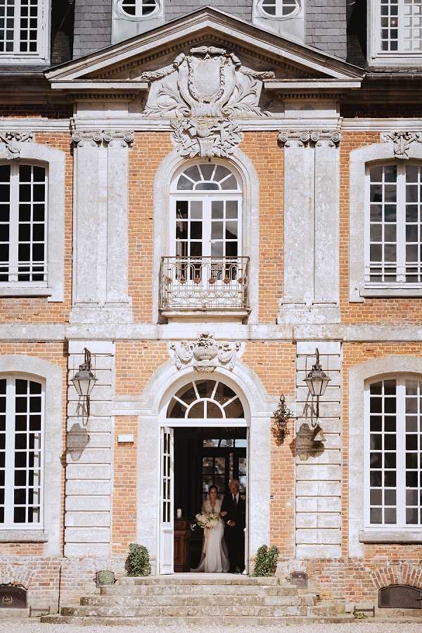 A bride in a white fitted gown holding a large ivory and white bouquet stands in the arched doorway of a French chateau alongside a male companion, appearing to be making her entrance or exit from the building. The shot is a wide portrait framing the full facade of the chateau, which features warm terracotta brick, ornate white stone quoins, carved decorative crests above the doorway and central window, wrought iron wall lanterns flanking the entrance, and a Juliet balcony with an iron railing on the upper level. Small boxwood topiaries flank the base of the entrance steps. The two figures are relatively small within the frame, emphasizing the scale and classical French architecture of the building. Potential venue feature image.