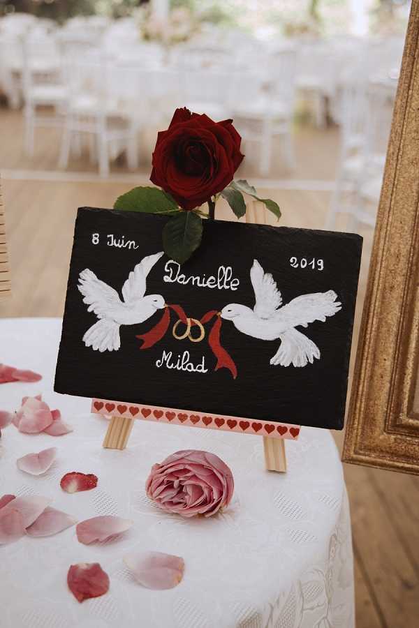Close-up detail shot of a welcome sign display at a wedding reception held indoors in a bright, wood-floored venue. The sign is a painted slate board propped on a small wooden easel, featuring two hand-painted white doves holding gold rings tied with a deep red ribbon, with the couple's names 'Danielle & Milad' and the date '8 Juin 2019' written in white script. A single deep red rose is tucked behind the top of the slate, and the surface below is scattered with dusty pink rose petals alongside a small blush garden rose. A gold ornate mirror frame is partially visible on the right, and white chair and round table settings are softly blurred in the background reception space.